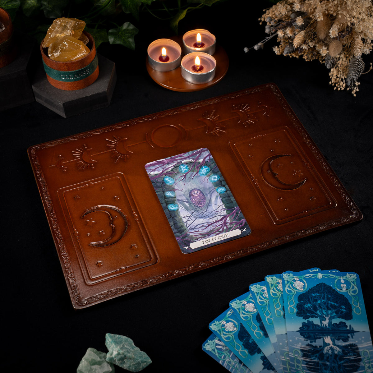 The Astro handmade leather tarot mat with a single tarot card on the centre placement, beside lit candles, crystals and a fanned deck