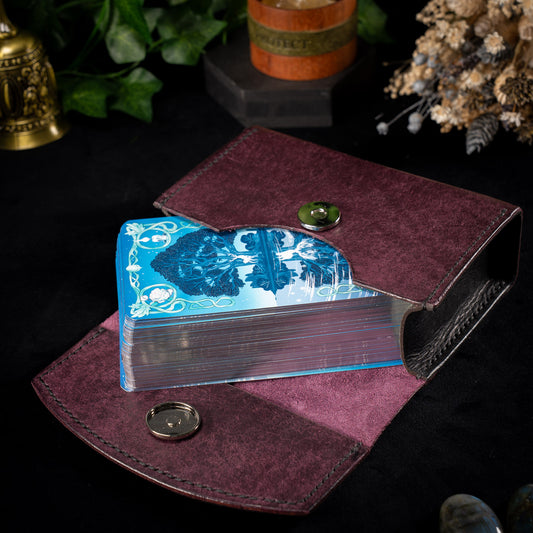 The Oracle handmade leather tarot deck case in plum, open with a gilt edged tarot deck lifting out of the curved front recess