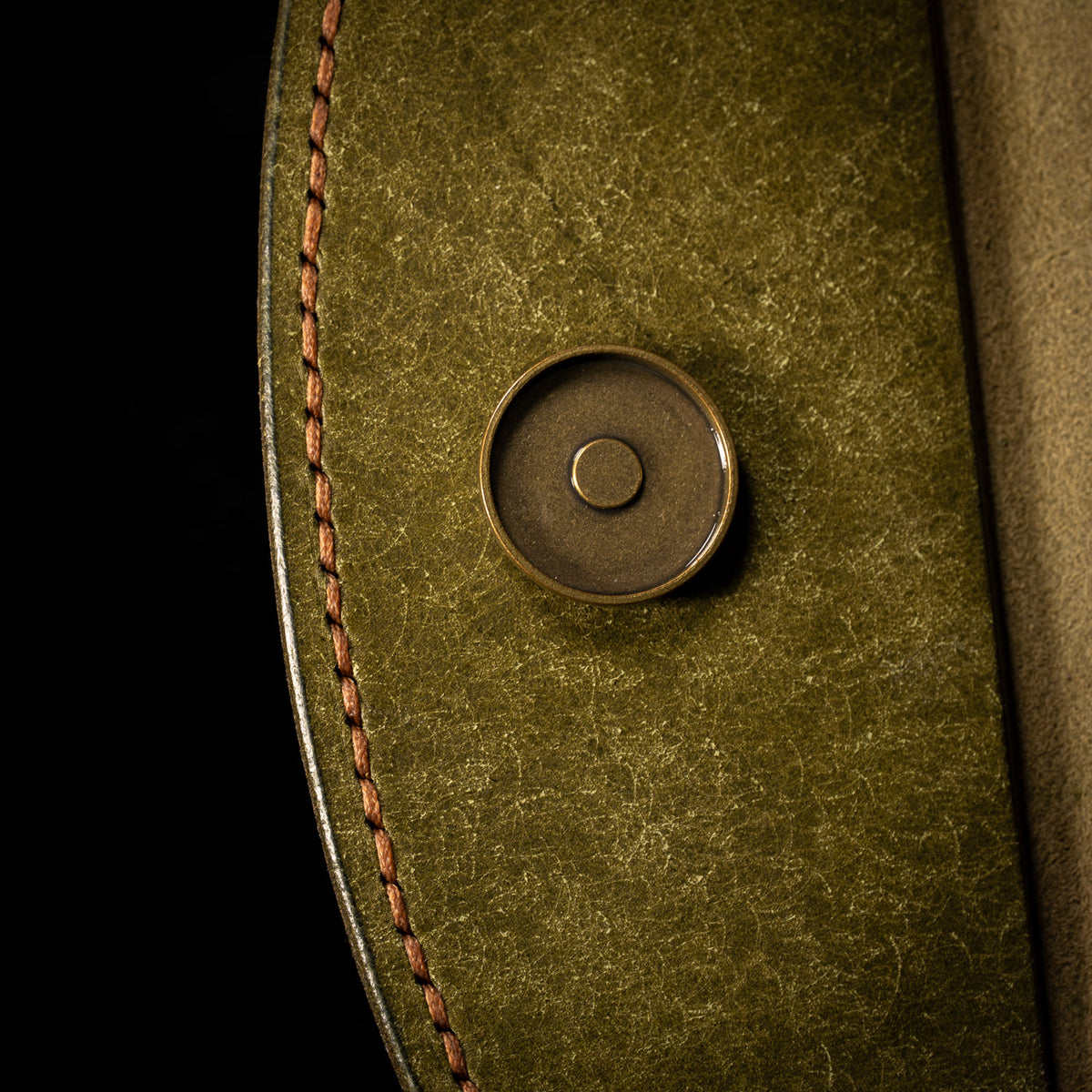 Close up of the brass magnetic snap and hand stitched tan thread running along the burnished edge of the olive flap